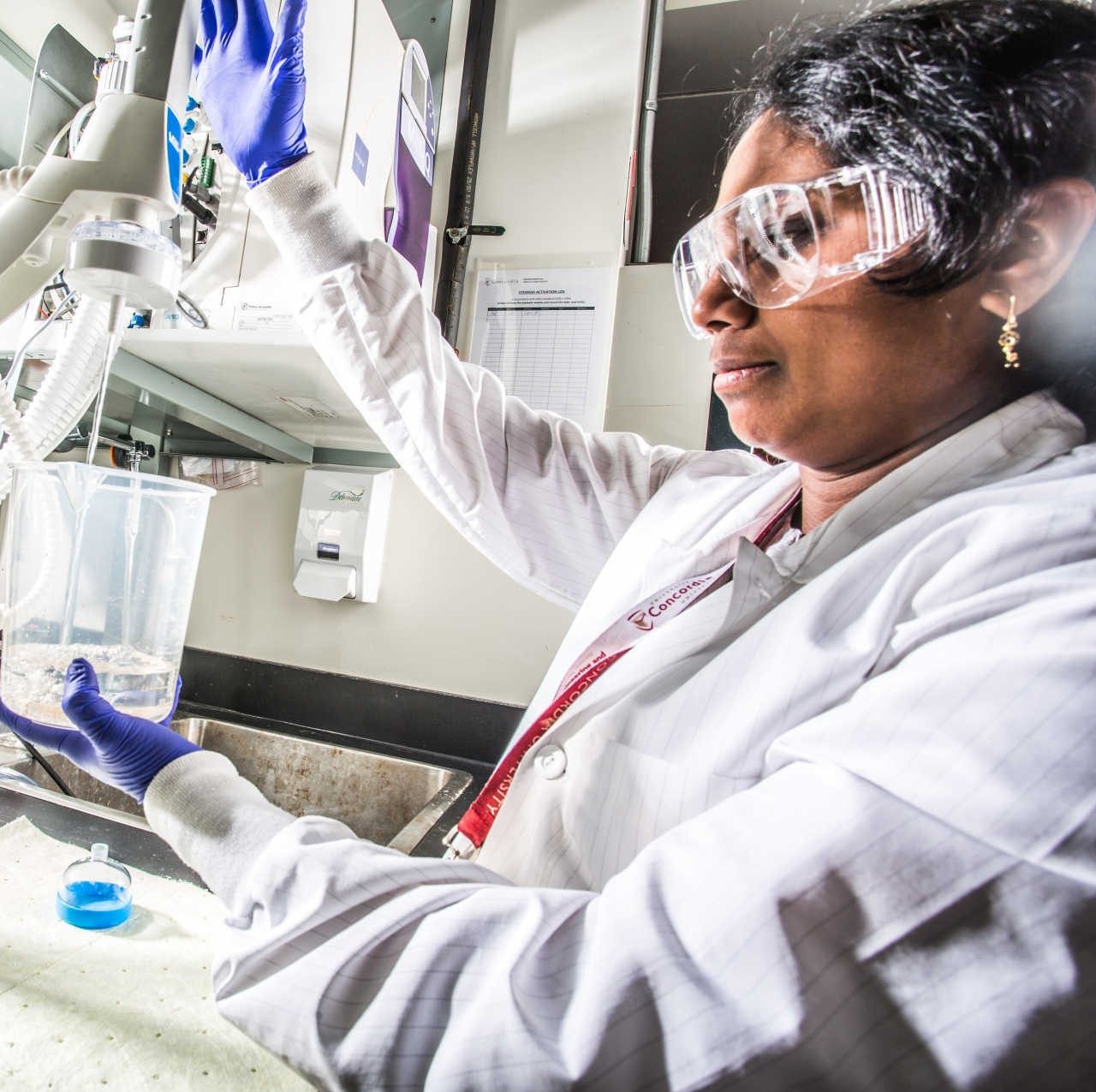 A woman in lab gear and a lab setting uses a mixer to stir a beaker with fluid in it. A woman in lab gear and a lab setting uses a mixer to stir a beaker with fluid in it.
