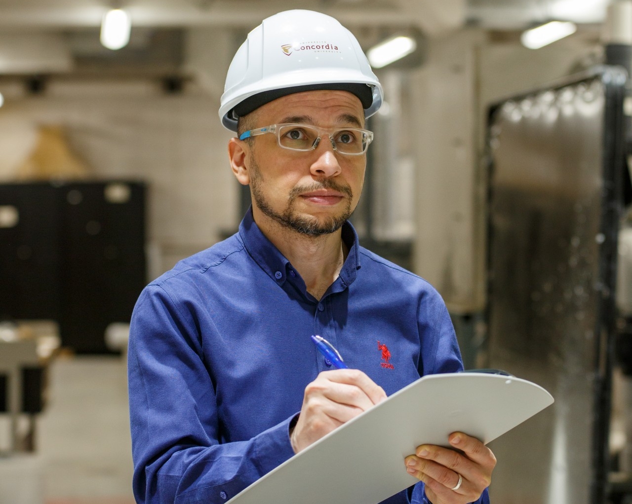 A person wearing a hard hat and safety goggles and holding a clipboard moves through an industrial area where several machines and pieces of equipment are located.