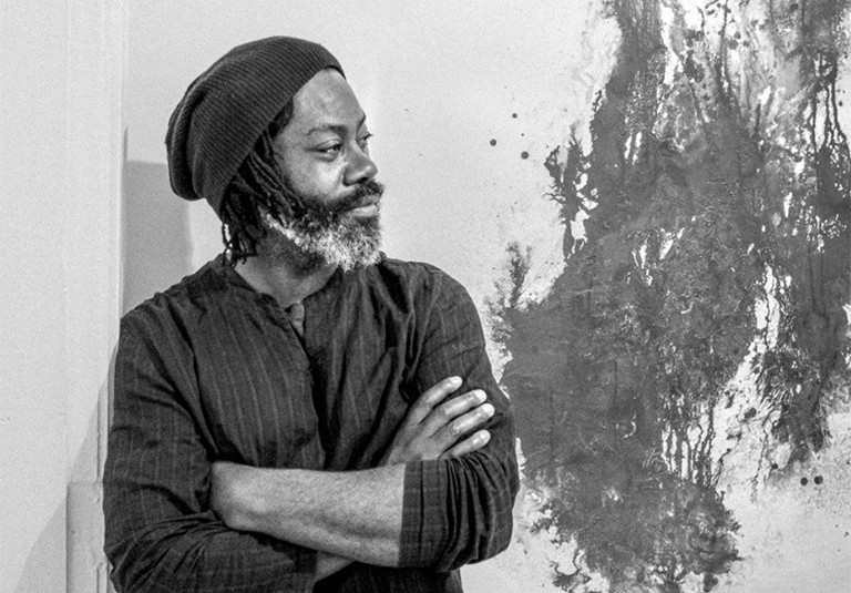 Profile of a man with a beard, wearing a woollen beanie, a striped top, standing beside an abstract painted artwork.
