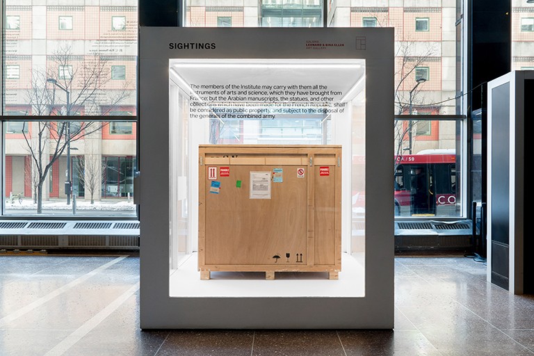 An exhibition art piece: A large square glass cube with a wooden crate inside it, in a public interior space.